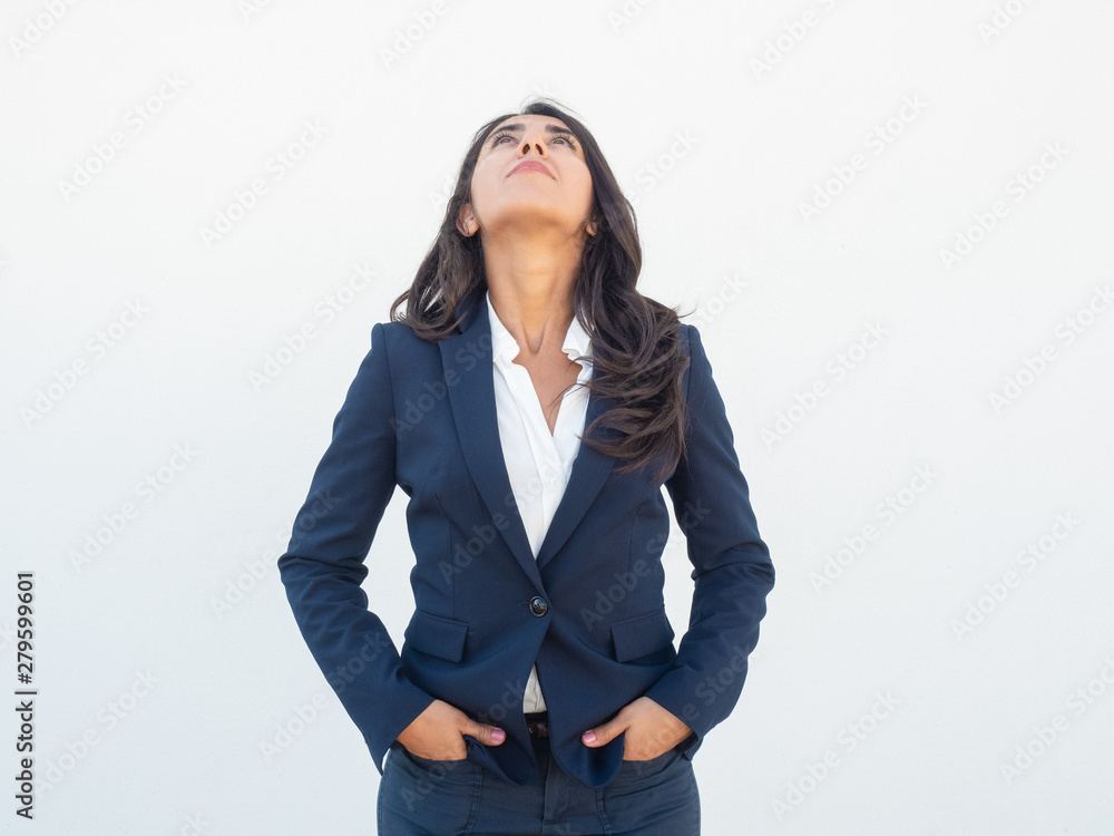 Serious positive businesswoman looking up. Front view of young Latin woman in office suit keeping hands in pocket and turning face up. Looking up concept Serious positive businesswoman looking up. Front view of young Latin woman in office suit keeping hands in pocket and turning face up. Looking up concept