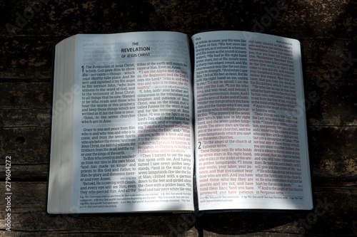 Open Bible in Revelation of Jesus Christ in chapters 1 and 2 with sunshine in the texts. Background with wooden table.