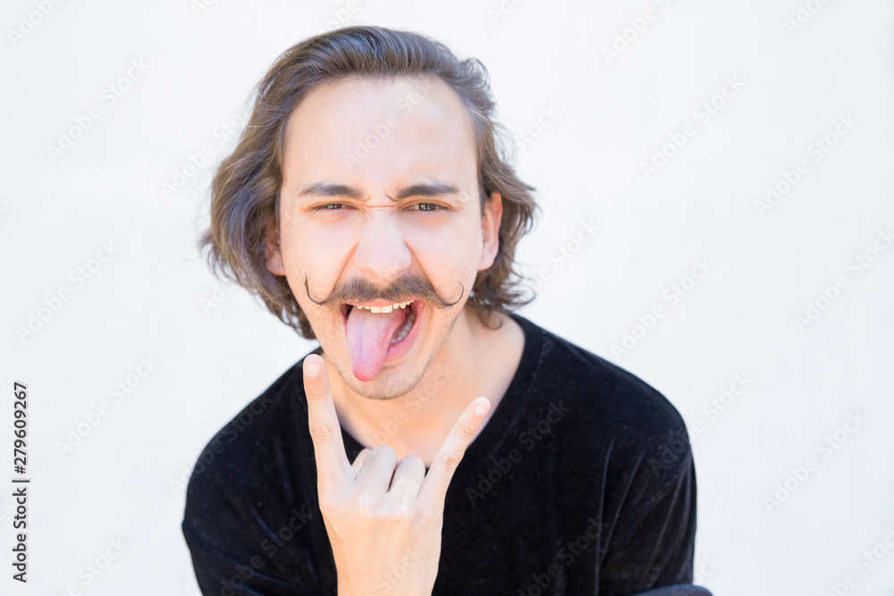 Handsome young man with mustache gesturing. Stylish guy showing tongue and devil horns sign. Rock sign concept Handsome young man with mustache gesturing. Stylish guy showing tongue and devil horns sign. Rock sign concept