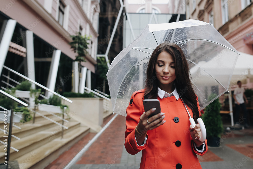 Fototapeta premium Beautiful woman with umbrella rainy day in red coat dialing number on phone making video call using smart phone and standing near old building.