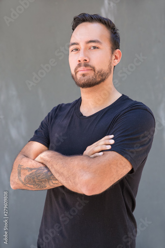 Serious bearded man looking at camera. Handsome confident tattooed man standing with crossed arms and looking at camera on grey background. Confidence concept