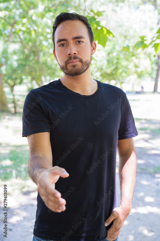 Young man reaching hand and looking at camera. Serious bearded man standing ready for handshake outdoor. Gesture concept Young man reaching hand and looking at camera. Serious bearded man standing ready for handshake outdoor. Gesture concept