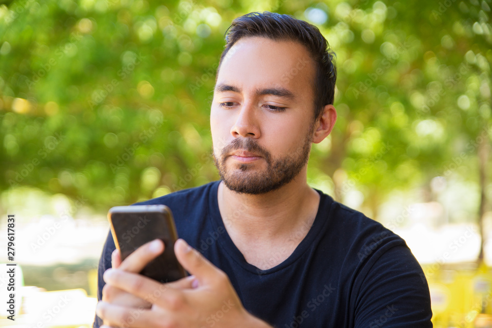 Serious man using mobile phone in park. Handsome bearded young man in black t-shirt using smartphone outdoor. Technology concept Serious man using mobile phone in park. Handsome bearded young man in black t-shirt using smartphone outdoor. Technology concept