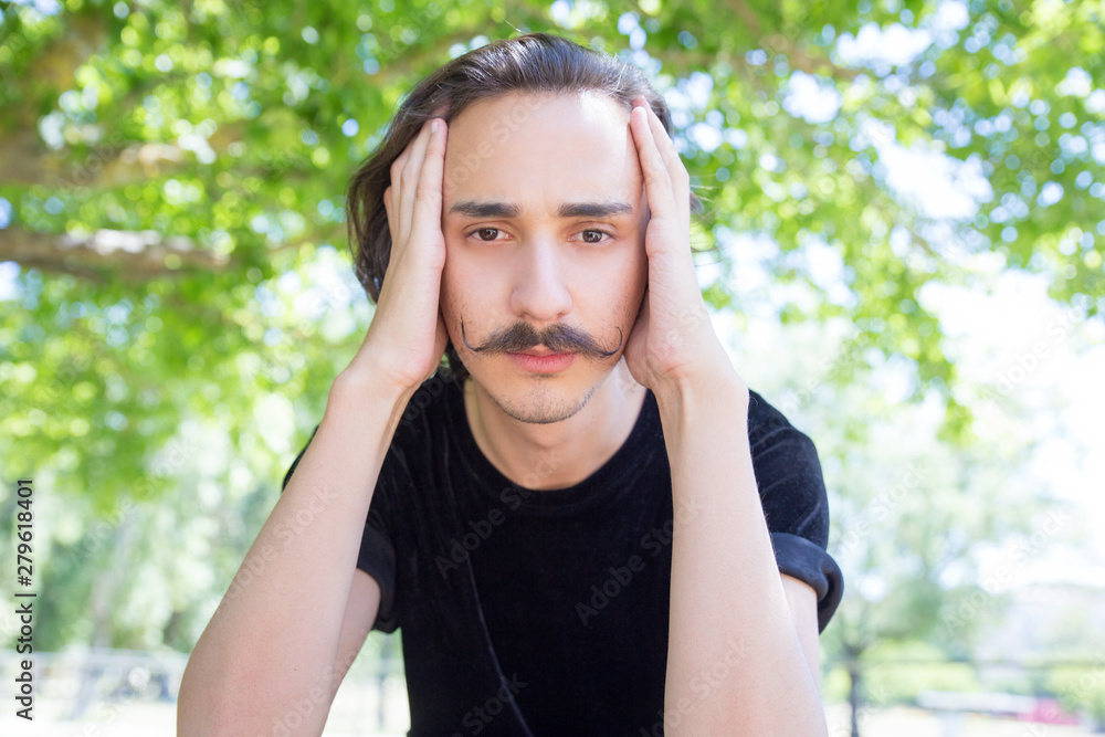 Tired man with handlebar mustache resting in park. Brunet looking at camera while sitting in park and covering ears with hands. Concept of fatigue
