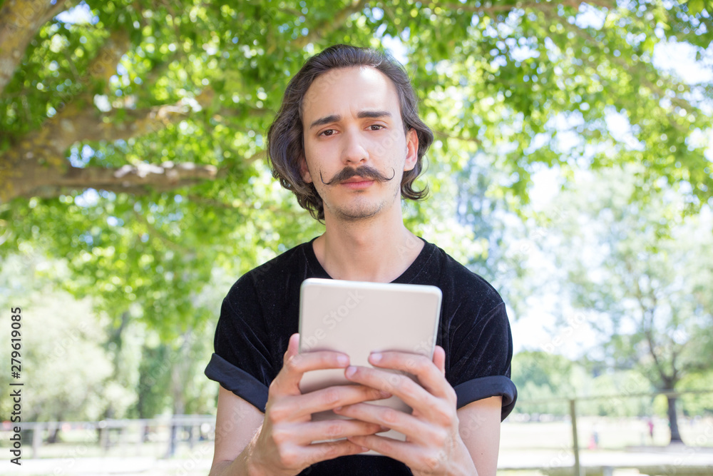Front view of serious man with tablet looking at camera. Thoughtful young brunet with tablet posing in park. Technology concept Front view of serious man with tablet looking at camera. Thoughtful young brunet with tablet posing in park. Technology concept