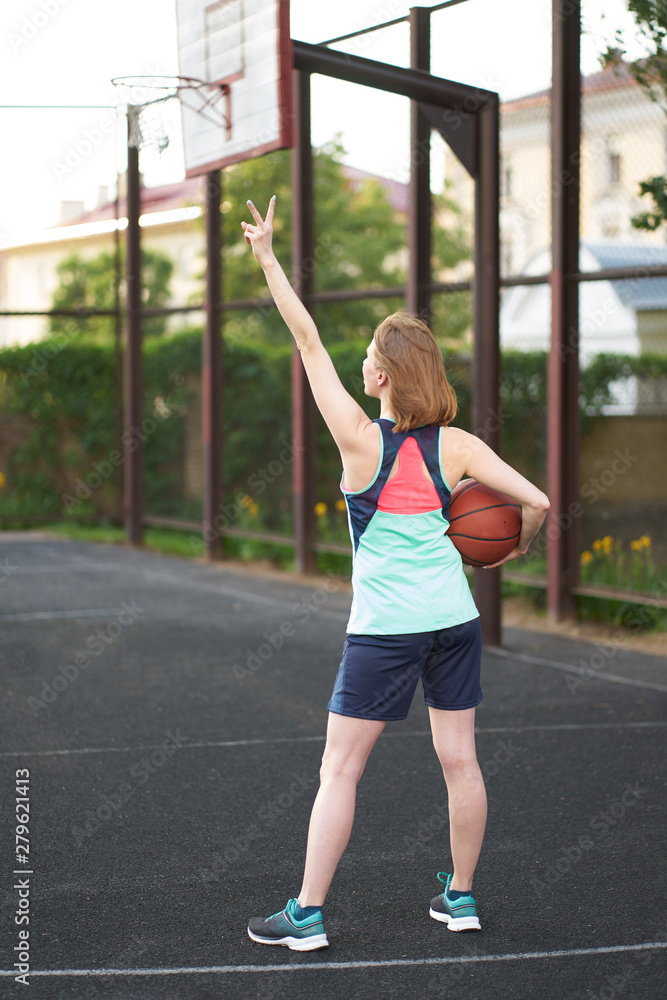 Redhead smiling caucasian girl shows victory sign with her fingers at outdoor street basketball court Redhead smiling caucasian girl shows victory sign with her fingers at outdoor street basketball court