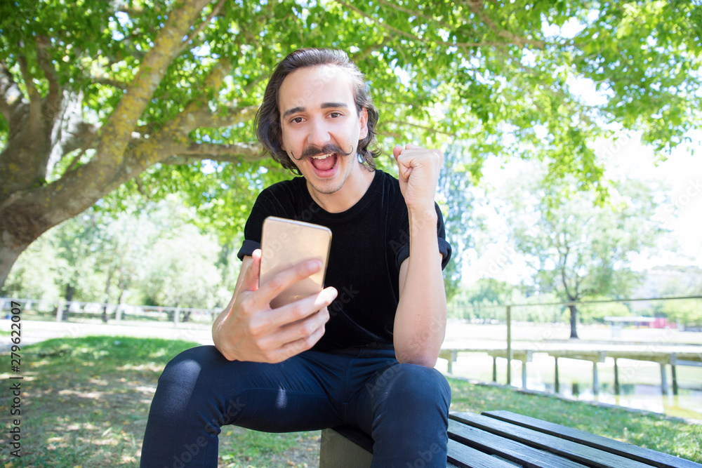 Successful young guy with smartphone looking at camera. Stylish guy with mustache holding raised fist. Technology concept Successful young guy with smartphone looking at camera. Stylish guy with mustache holding raised fist. Technology concept
