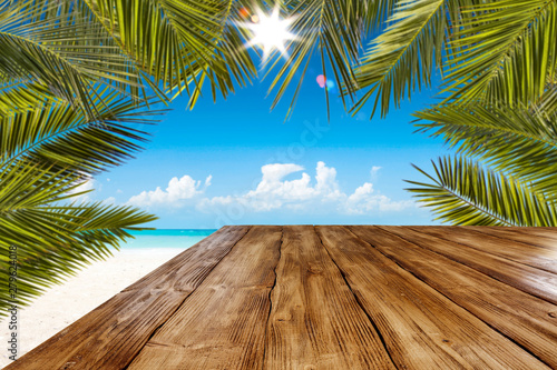 Table background with wooden board in the beautiful ocean and beach view. Palm leaves and clear sunny sky. Free space on a wooden table for an advertising product.