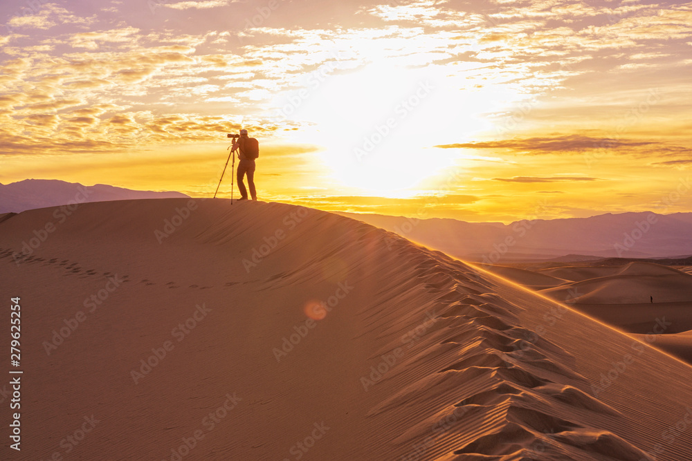 Photographer behind tripod standing with camera on sand dune silhouetted by sunrise in desert, Death Valley California