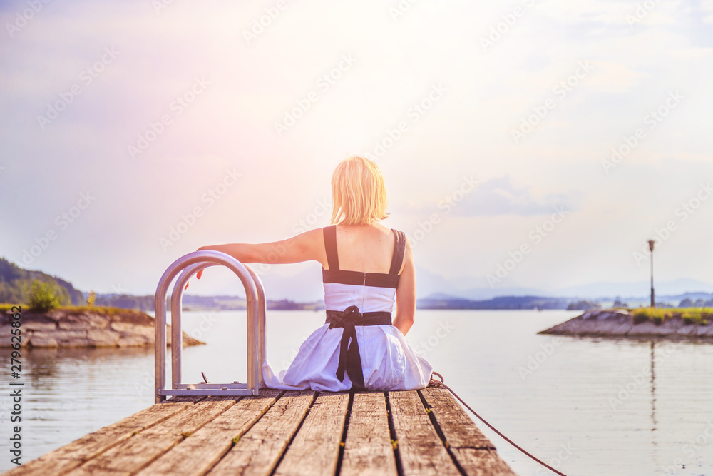 Beautiful young woman with white dress is sitting on a footbridge and enjoying the view, summer time