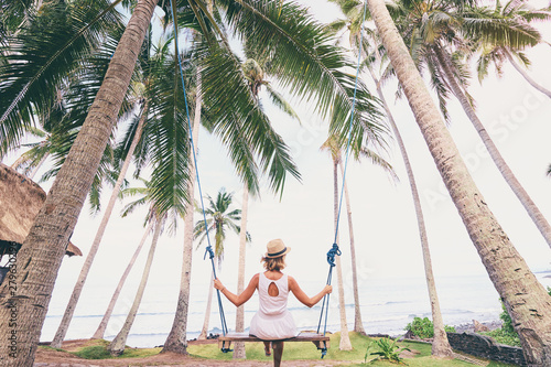 Vacation concept. Happy young woman in white dress and hat swinging at palm grove enjoying sea view. photo