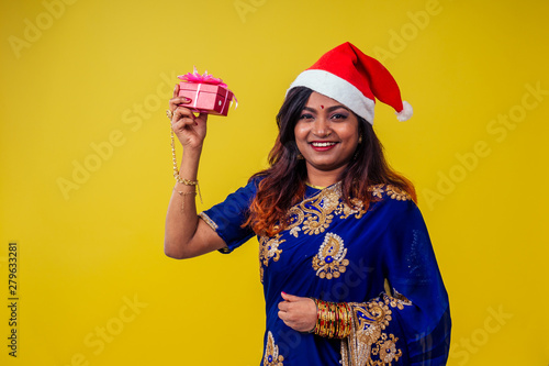 woman portrait wearing traditional india sari and Christmas Santa hat hold christmas gifton yellow background studio photo