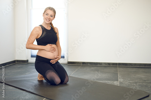 Happy pregnant young woman doing exercises kneeling on a yoga mat clasping her swollen belly in both hands while smiling at the camera