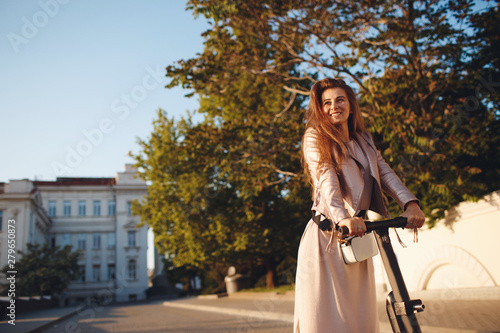 Portrait of a smiling pretty girl in pink clothes, rides on a scooter and looks at the camera, background city in the sunset. Walk on a kick scooter.Beautiful woman riding a scooter, walking, leisure