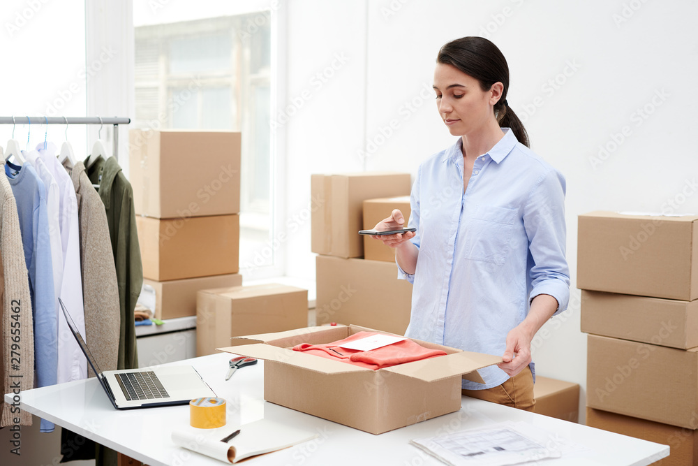 Young female shop-assistant photographing parcel with folded casualwear Young female shop-assistant photographing parcel with folded casualwear