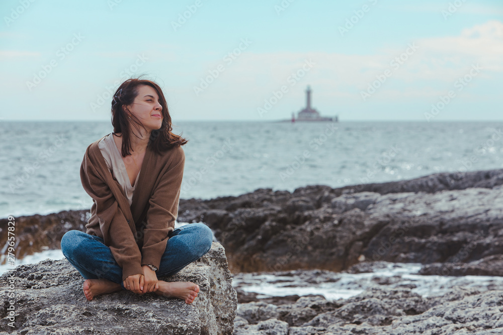 woman sitting by rocky sea beach in wet jeans lighthouse on background