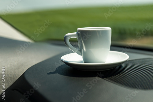 White coffee Cup with coffee stands on the black panel of the car. Through the windshield you can see the green field. Copy space. Concept: auto travel with a drink.