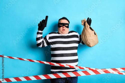 fat burger with raised arms holding a sack with money and pistol and looking at the camera. isolated blue background, warning tape in the foreground photo