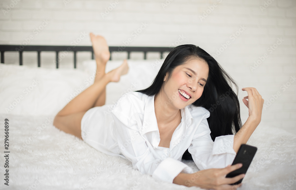 Beautiful young woman in white shirt using video call or self portrait selfie from smartphone on white bed in bedroom, Happy woman relaxing, Smiles and laughs, Communication and social network-Image Beautiful young woman in white shirt using video call or self portrait selfie from smartphone on white bed in bedroom, Happy woman relaxing, Smiles and laughs, Communication and social network-Image