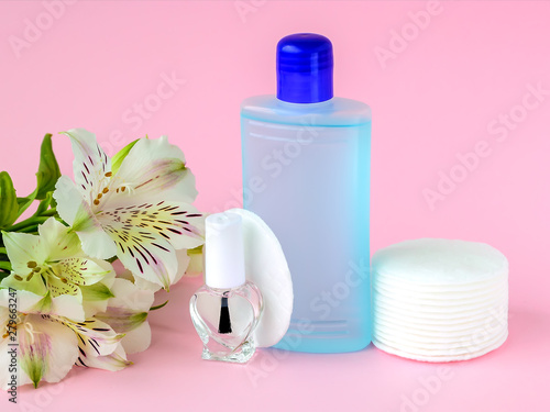 Glass bottle with colorless nail polish, plastic bottle with nail varnish remover, cotton pads and white flowers on a pastel pink background. Manicure, pedicure, nail care products.