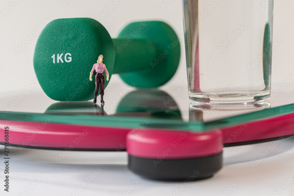 Miniature woman figure standing on the digital electronic bathroom scale for weight of human body. Glass of water and green dumbbells at shallow depth of field background. slimming concept Miniature woman figure standing on the digital electronic bathroom scale for weight of human body. Glass of water and green dumbbells at shallow depth of field background. slimming concept