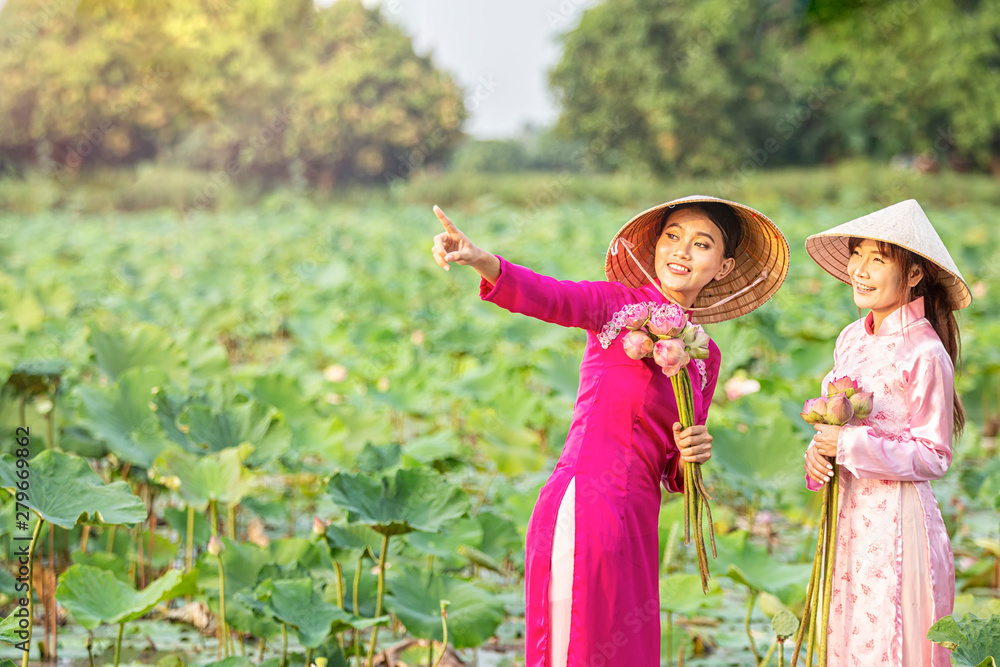 Obraz premium Vietnamese girls are inviting friends to look at the lotus flowers. Vietnamese dress with pink lotus flowers. Female boating on lakes harvest water lilies.