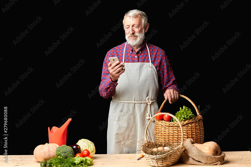 Senior village man texting message to his wife on smartphone, looking anxious about her being late, going to cook dinner for two, bought in organic farmer's shop lots of fresh vegetables and eggs. Senior village man texting message to his wife on smartphone, looking anxious about her being late, going to cook dinner for two, bought in organic farmer's shop lots of fresh vegetables and eggs.