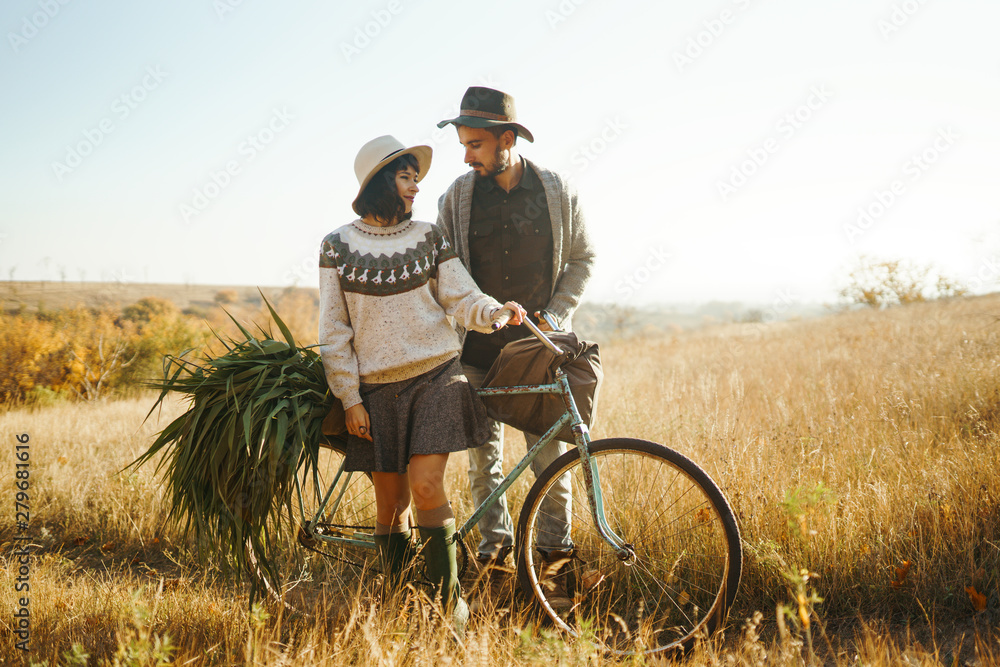 Lovely hipster couple with bike . Couple wearing beautiful hats and sweaters. Lifestyle, happy couple of two play on a sunny day in the park. The concept of youth, love and lifestyle. Sunset in autumn - obrazy, fototapety, plakaty 