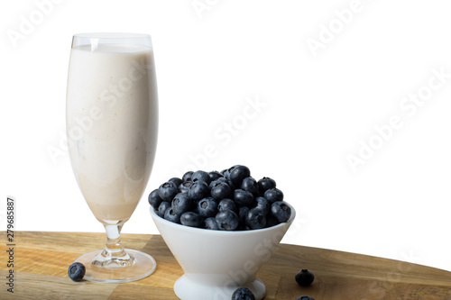 Blueberries in a white bowl and scattered around a glass of milkshake on a wooden board on a white background isolated