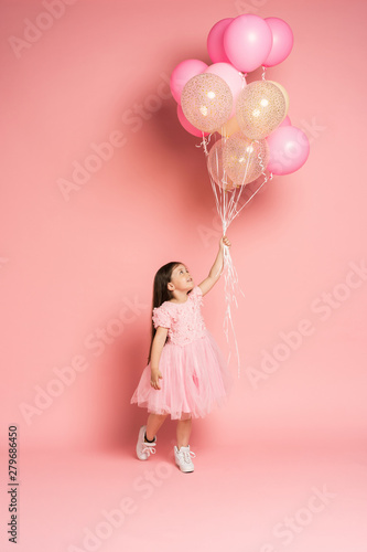 Happy celebration of birthday party with flying balloons of charming cute little girl in tulle dress smiling to camera isolated on pink background. Charming smile  expressing happiness