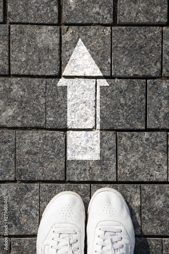 feet white sneakers standing on a cobblestone pavement in front the white direction sign to go ahead.