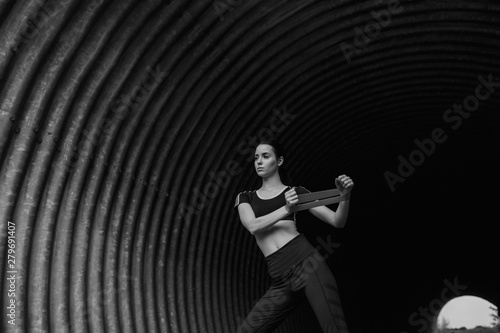 Sports woman on tunnel urban background. Fitness model working out outdoor. Young beautiful slim brunette girl doing stretching warmup exercise with resistance band. Black and white.