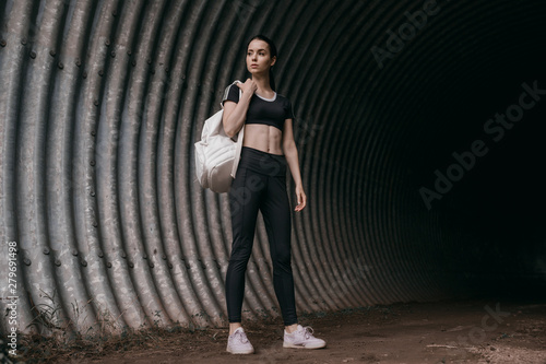 Sports woman posing in fashion sportswear on tunnel urban gray background. Fitness model working out outdoor. Beautiful slim girl in trendy black leggings, top and white backpack. © KDdesignphoto