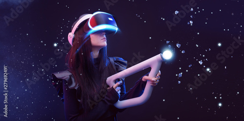 Beautiful woman in futuristic dress over dark magic light background. Gamer girl in glasses of virtual reality with controller in hands. Augmented reality, game, hobby concept. VR.