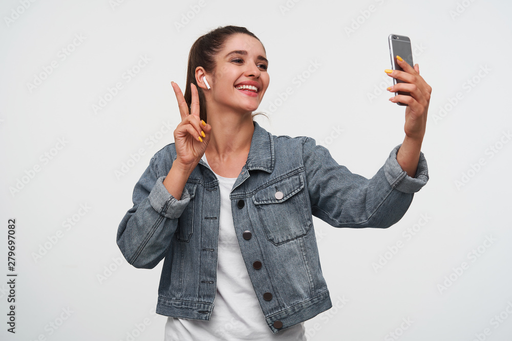 Young cheerful brunette lady wears in white t-shirt and denim jackets, holds smartphone and makes selfie with peace gesture. Stands over white background. Young cheerful brunette lady wears in white t-shirt and denim jackets, holds smartphone and makes selfie with peace gesture. Stands over white background.