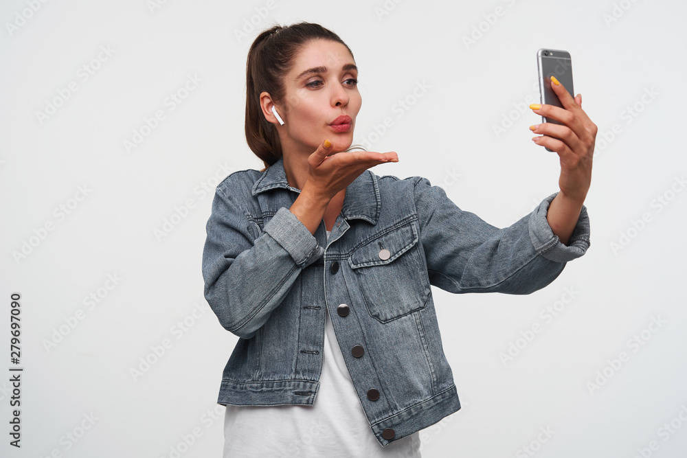 Young happy brunette woman wears in white t-shirt and denim jackets, holds smartphone and sends kiss to the videochat. Stands over white background. Young happy brunette woman wears in white t-shirt and denim jackets, holds smartphone and sends kiss to the videochat. Stands over white background.