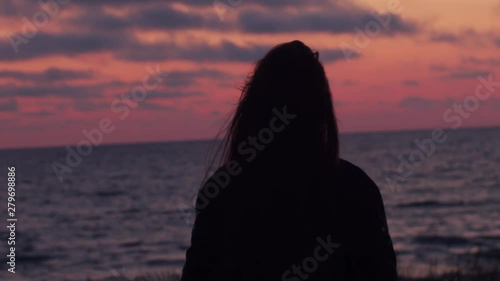 Lonely woman standing at seaside looking to sea landscape in evening natural light. Back view of beautiful girl stroking hair backlit by setting rising sun slow motion. female beauty hair care