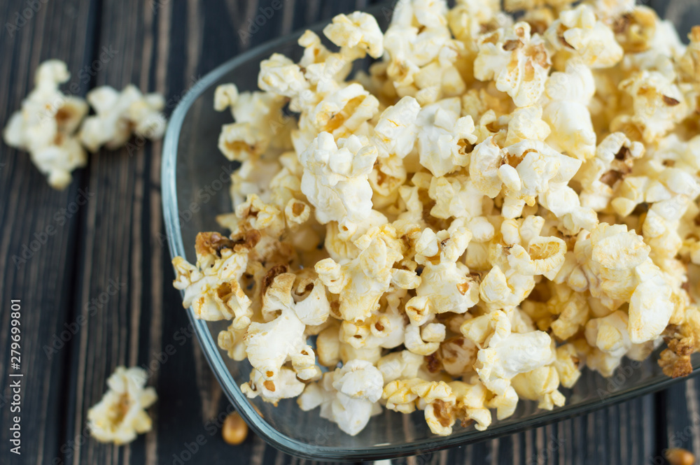 Homemade vegan fresh crunchy salted popcorn in a glass plate on a dark wooden table. A useful snack for a movie, an idea for a meal. Dark mood close-up top view. Homemade vegan fresh crunchy salted popcorn in a glass plate on a dark wooden table. A useful snack for a movie, an idea for a meal. Dark mood close-up top view.