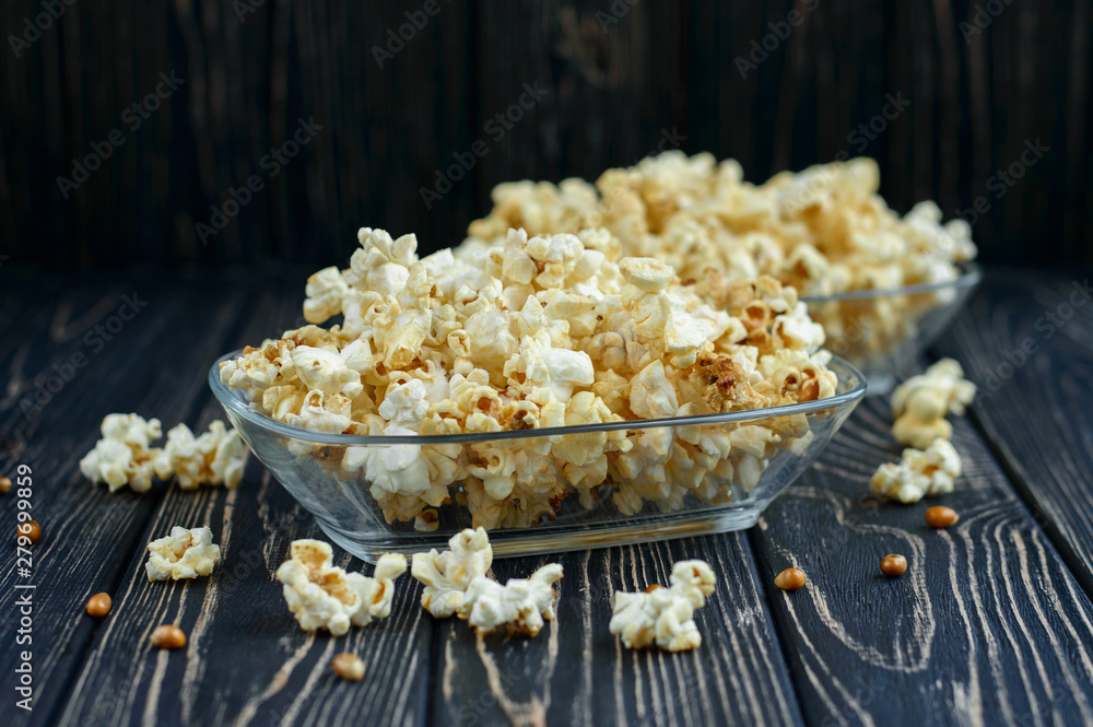 Homemade vegan fresh crunchy salted popcorn in a glass plate on a dark wooden table. A useful snack for a movie, an idea for a meal. Dark mood. Homemade vegan fresh crunchy salted popcorn in a glass plate on a dark wooden table. A useful snack for a movie, an idea for a meal. Dark mood.
