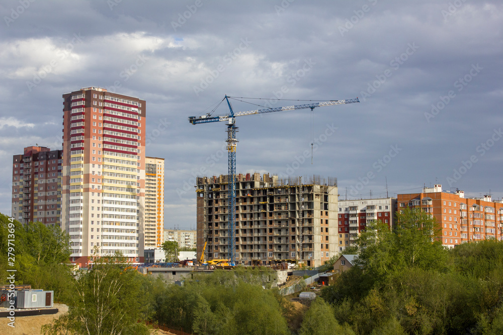 tall tower crane near a multi-storey modern house under construction near a green forest against the background of finished houses under a cloudy sky tall tower crane near a multi-storey modern house under construction near a green forest against the background of finished houses under a cloudy sky