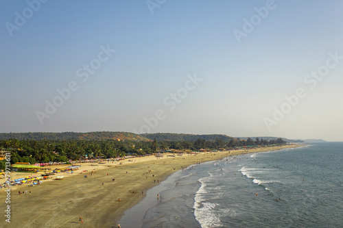 Arambol, Goa/India - 04.01.2019: people walk along the sandy beach of the sea coast and swim in the ocean against the backdrop of sun beds and palm jungle, aerial view