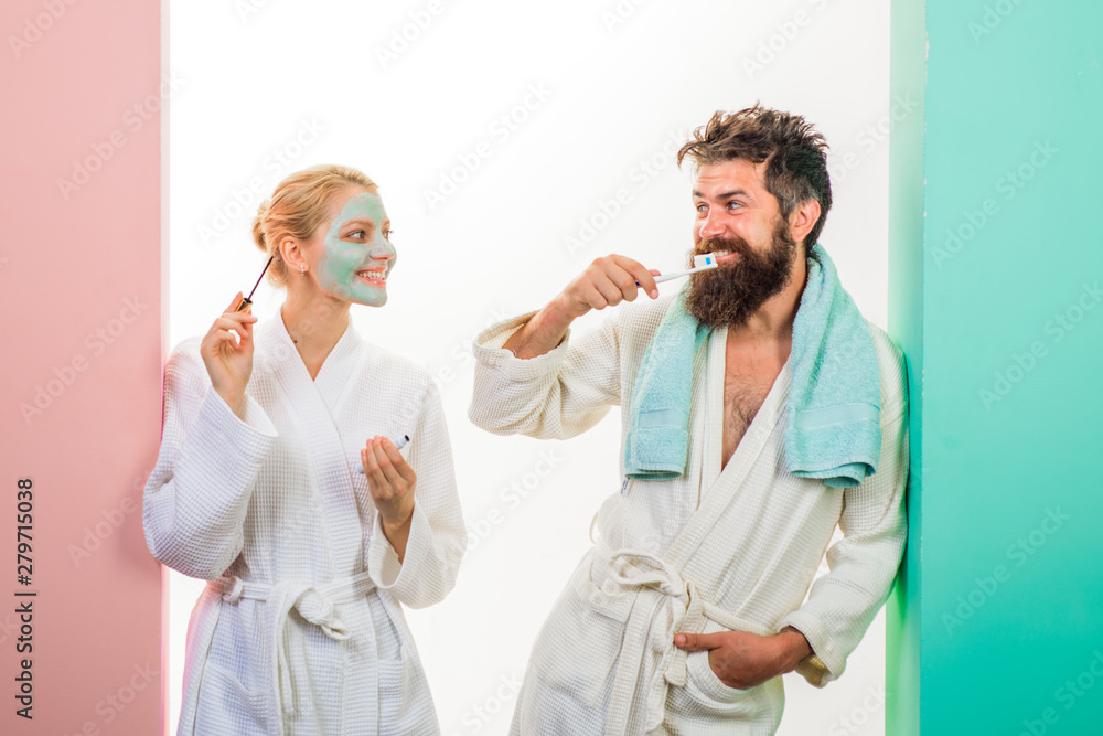 Obraz premium Morning preparation. Wife and husband preparing for work. Girl with cosmetic mask on face applying mascara. Couple. Woman applying mascara, man brushing teeth. Bearded man in bathrobe brushing teeth.