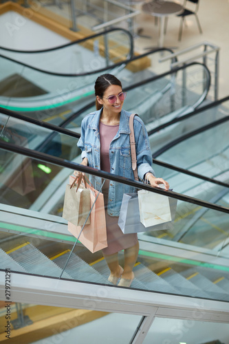 Positive young shopaholic in long jacket and dress moving up escalator and looking around mall