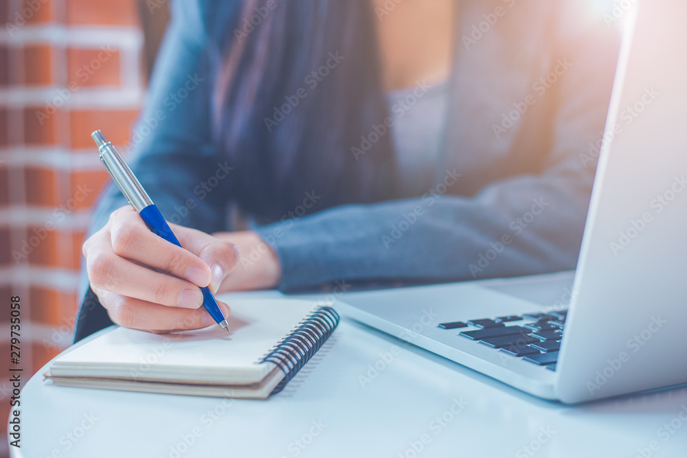 Business woman hand is writing on a notepad with a pen and have a laptop computer on the desk in the office.