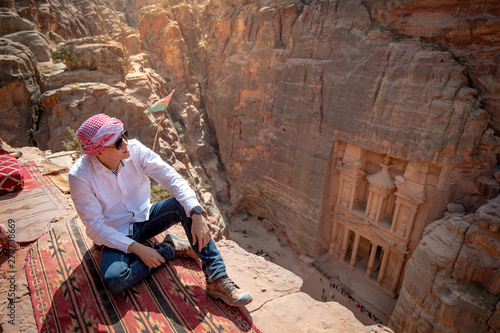 Asian man traveler sitting on carpet viewpoint in Petra ancient city looking at the Treasury or Al-khazneh, famous travel destination of Jordan and one of seven wonders. UNESCO World Heritage site.