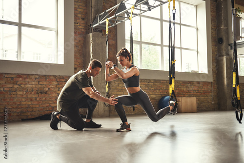 Obraz Robienie przysiadów. Pewny siebie osobisty trener pokazuje szczupłą wysportowaną kobietę, jak robić przysiady z paskami fitness Trx podczas treningu na siłowni.