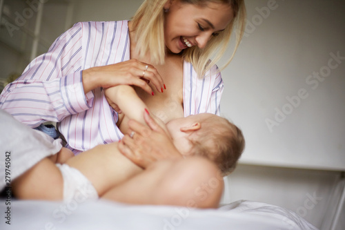 Indoor shot of charming cheerful young blonde woman in striped nightwear smiling broadly while breastfeeding her playful infant son who is biting nipple. Mother looking at baby, smiling happily