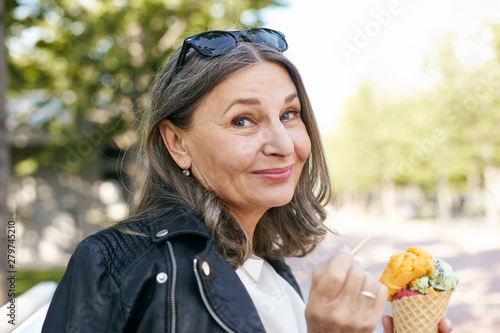 Stylish charming middle aged female in black leather jacket holding cone of colorful ice cream using wooden stick and smiling at camera, enjoying slow summer day, walking outdoors, feeling relaxed
