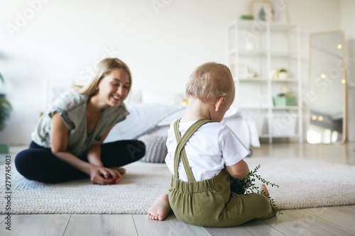 Beautiful positive young European mom sitting cross legged on floor with her infant son, watching him study plant. Blonde mother encouraging baby to do first steps in stylish bedroom interior