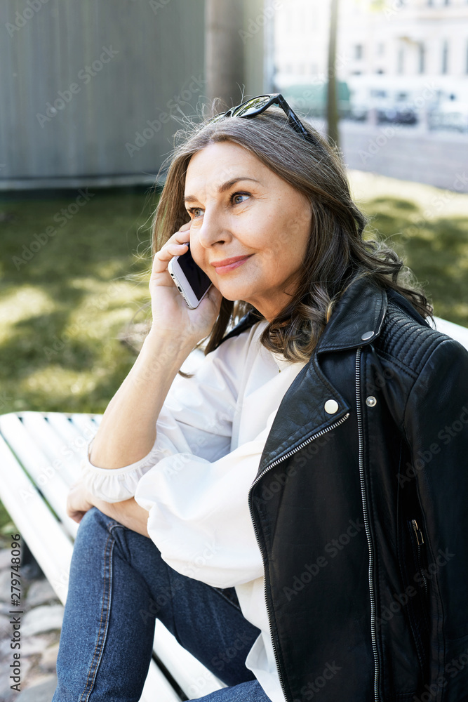 Vertical image of good looking modern middle aged female wearing stylish sunglasses, leather jacket and jeans calling taxi service using mobile. Energetic 50 year old talking on smart phone Vertical image of good looking modern middle aged female wearing stylish sunglasses, leather jacket and jeans calling taxi service using mobile. Energetic 50 year old talking on smart phone